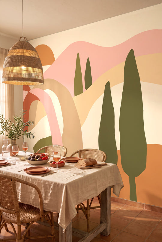 Salle à manger méditerranéenne avec table dressée et chaises en rotin devant un papier peint panoramique de collines toscanes terracotta et rose