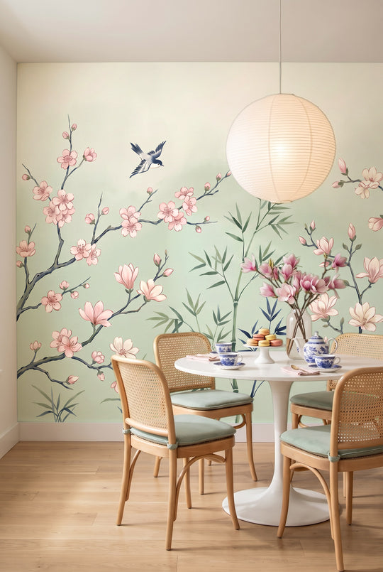Salle à manger avec table ronde blanche, service à thé et chaises cannées devant un papier peint chinoiserie de magnolias, bambous et oiseaux sur fond céladon