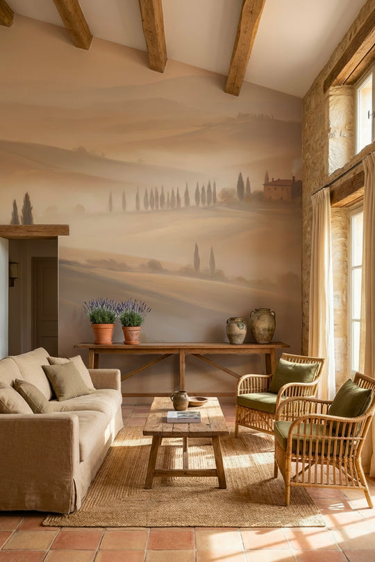 Salon méditerranéen avec canapé en lin, fauteuils en rotin et poutres apparentes devant un papier peint panoramique de collines toscanes dans la brume